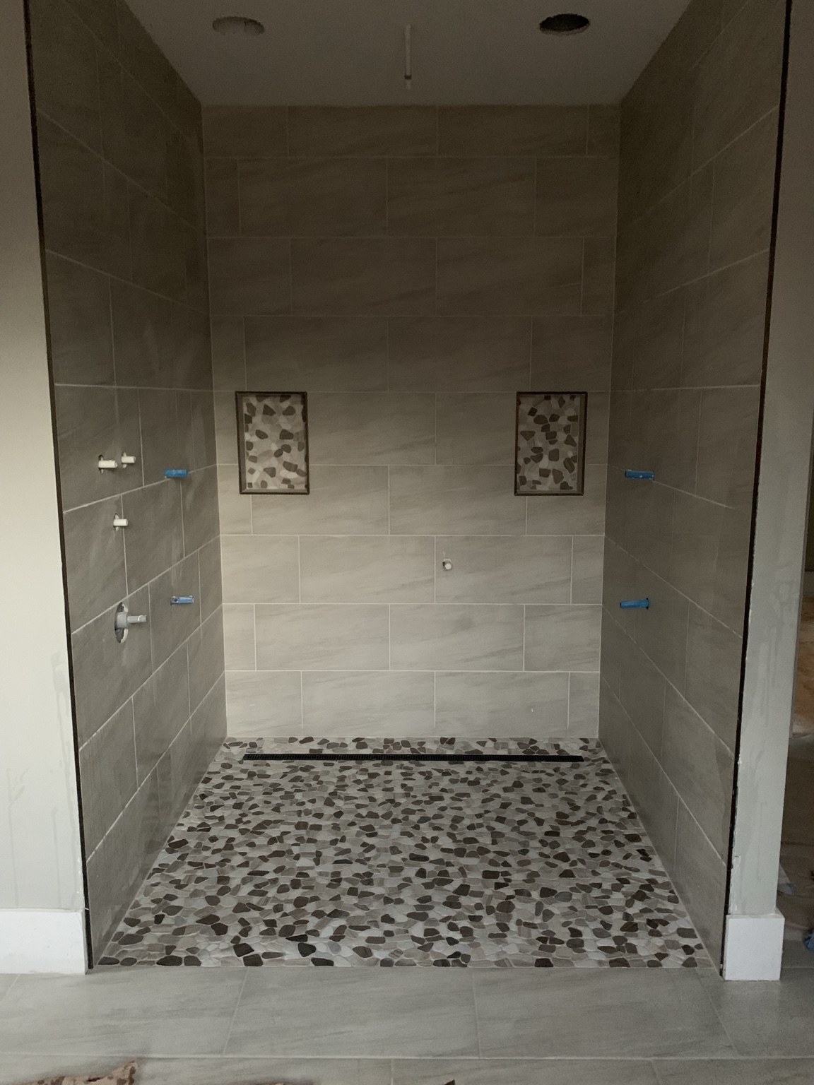 Large gray tiled shower with two pebble mosaic niches and a pebble floor during installation